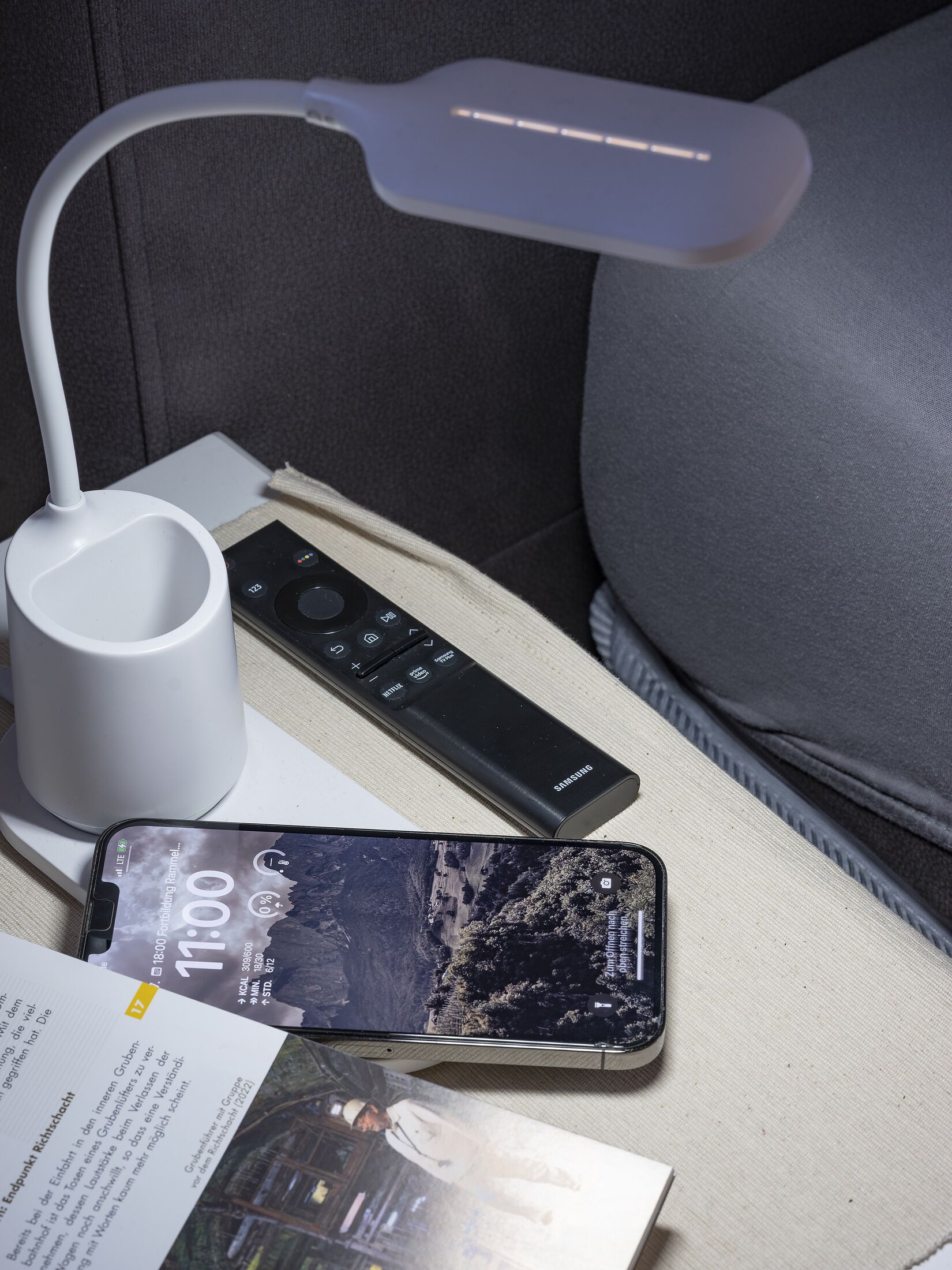 Ett litet bord vid sängen med en vit lampa, en fjärrkontroll, en smartphone och en broschyr.