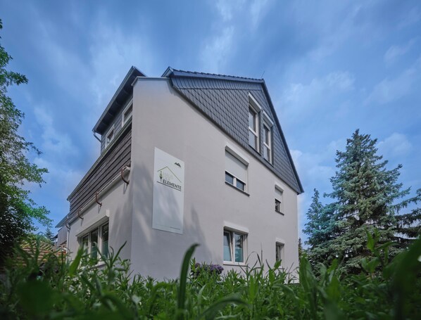 A two-story building with a sloped roof, a sign reading "HAUS DE ELEMENTE," and a garden in the foreground.