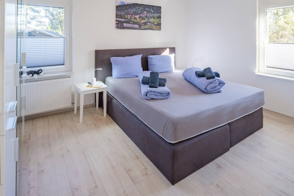 Ein Schlafzimmer mit Bett, Nachttisch, Lampe und einem Fenster mit Jalousien.