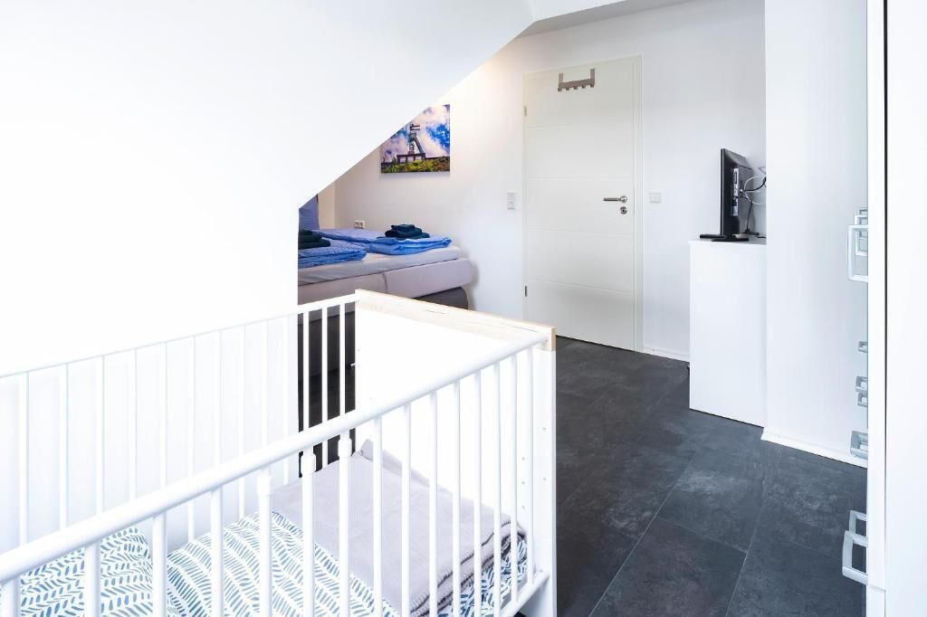 A modern room with a white crib, a bed with blue bedding, a white door, and a television on a white cabinet.