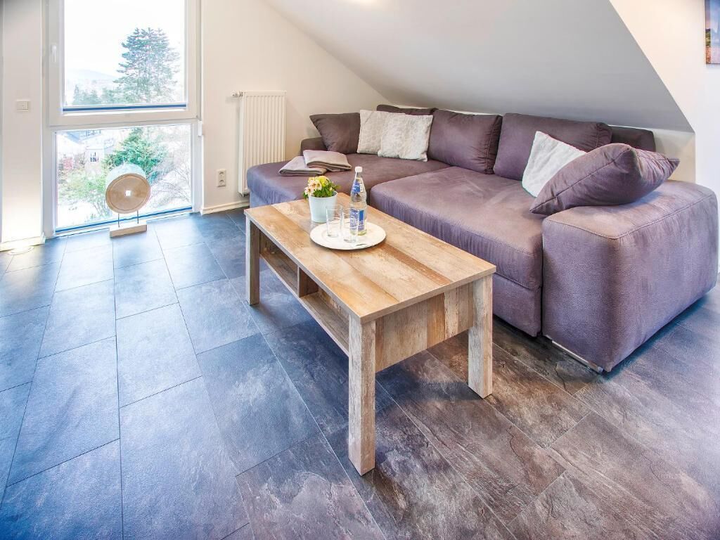 Un salon moderne avec un parquet en bois foncé, une table basse en bois, un canapé violet et une grande fenêtre laissant entrer la lumière naturelle.