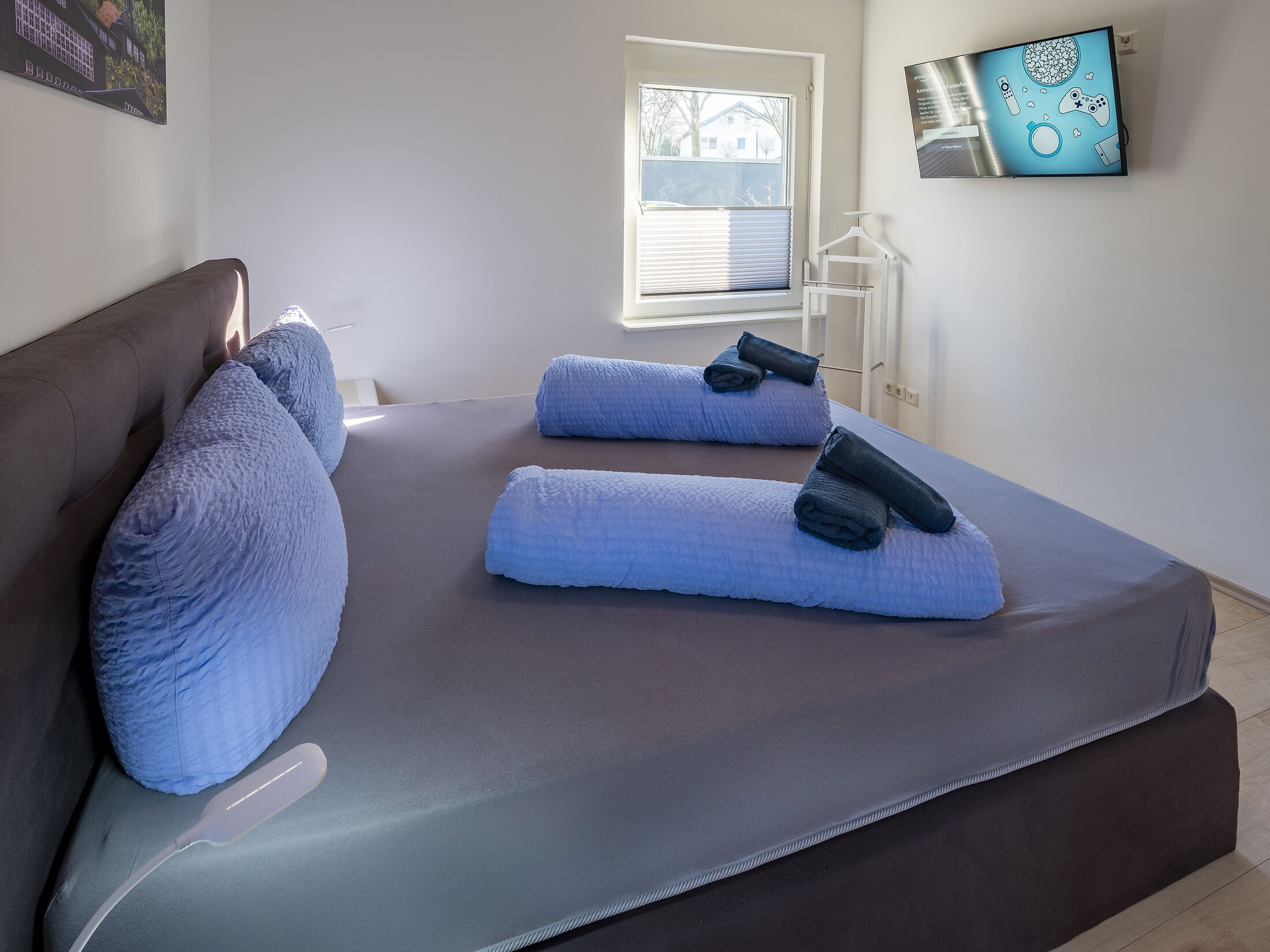 A bedroom with a large bed, two pillows, a bedside table, a window with blinds, a wall-mounted TV, and a framed picture.