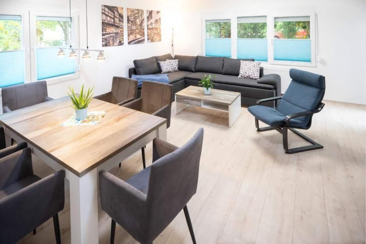 Ein modernes Wohnzimmer mit einem Holzesstisch, grauen Polsterstühlen, einem schwarzen Sofa und einem blauen Sessel.