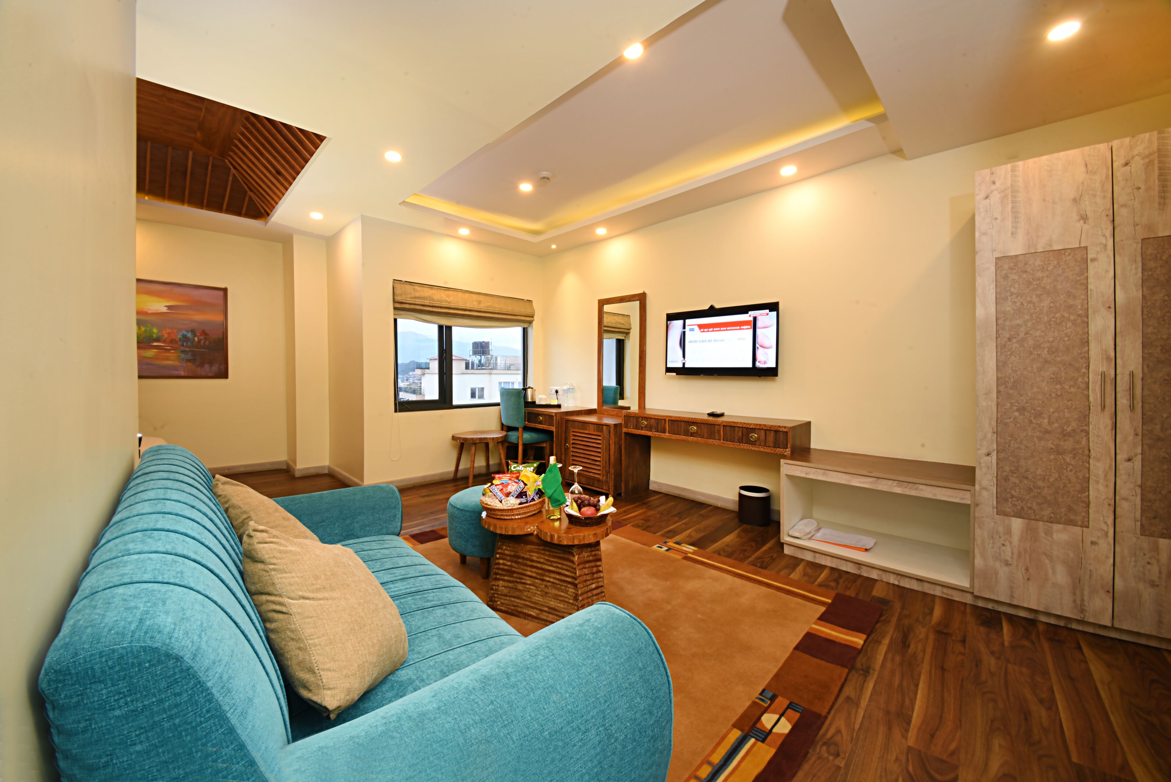 Un salón con sofá azul, piso de madera, televisor montado en la pared y vistas a la ciudad desde la ventana.