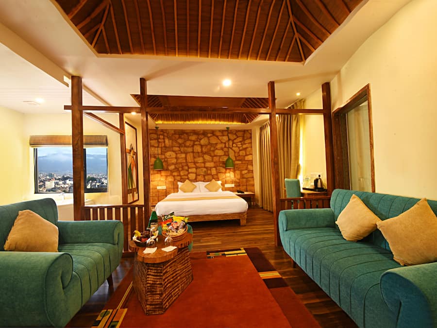 A hotel room with a stone wall, a large bed, two green sofas, a wooden ceiling, and a view of the city.
