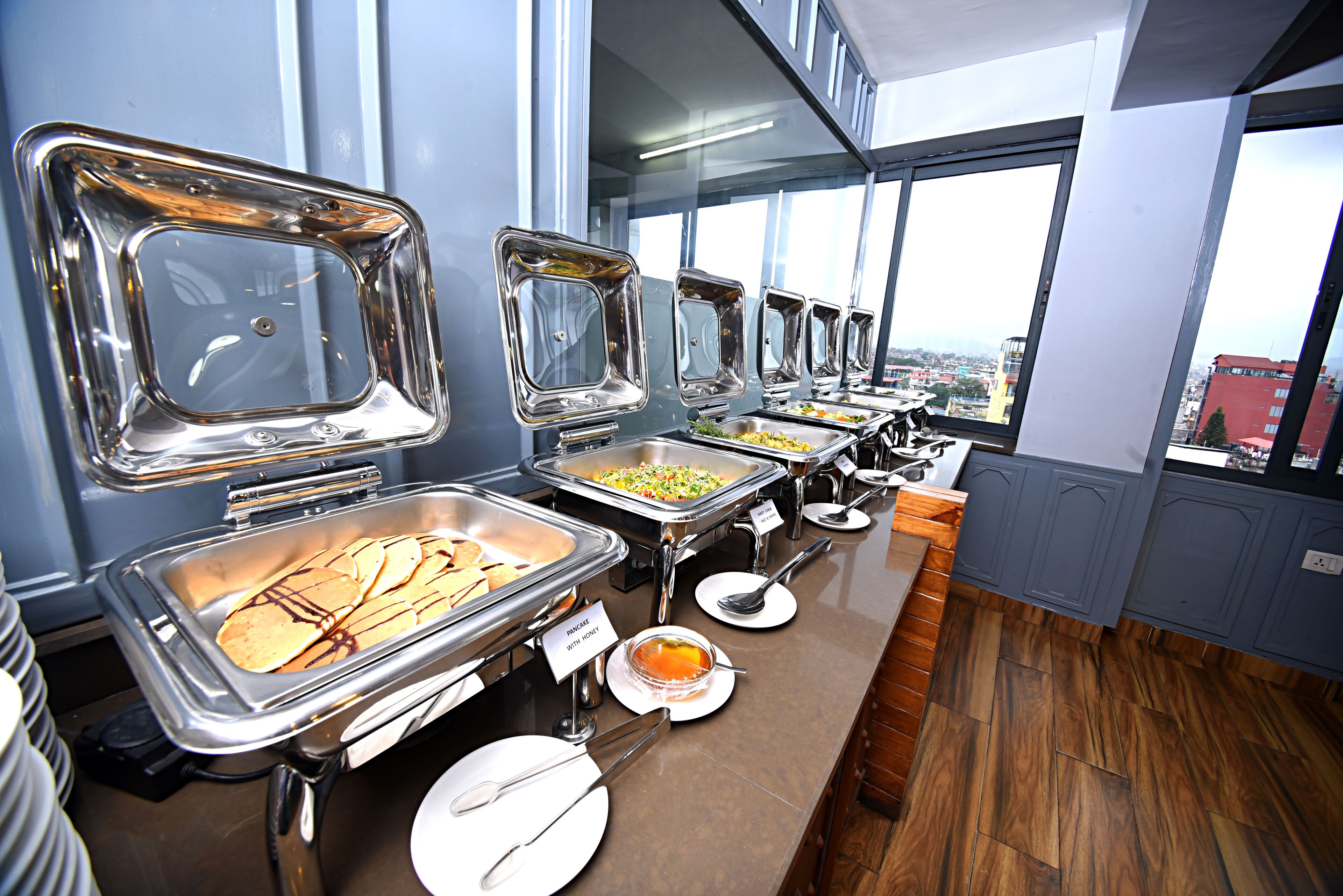 A buffet setup with multiple serving stations, including a pancake station and a soup station, in a modern kitchen with large windows.
