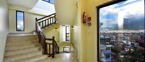 Eine Treppe mit hölzernem Geländer, das zu einer oberen Etage führt, ein großes Fenster mit Blick auf die Stadt und ein an der Wand befestigter Feuerlöscher.