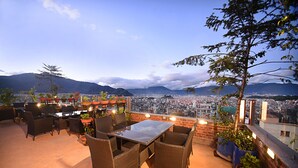 Un patio al aire libre con mobiliario de mimbre y plantas en macetas, con vistas a un paisaje urbano y montañas.