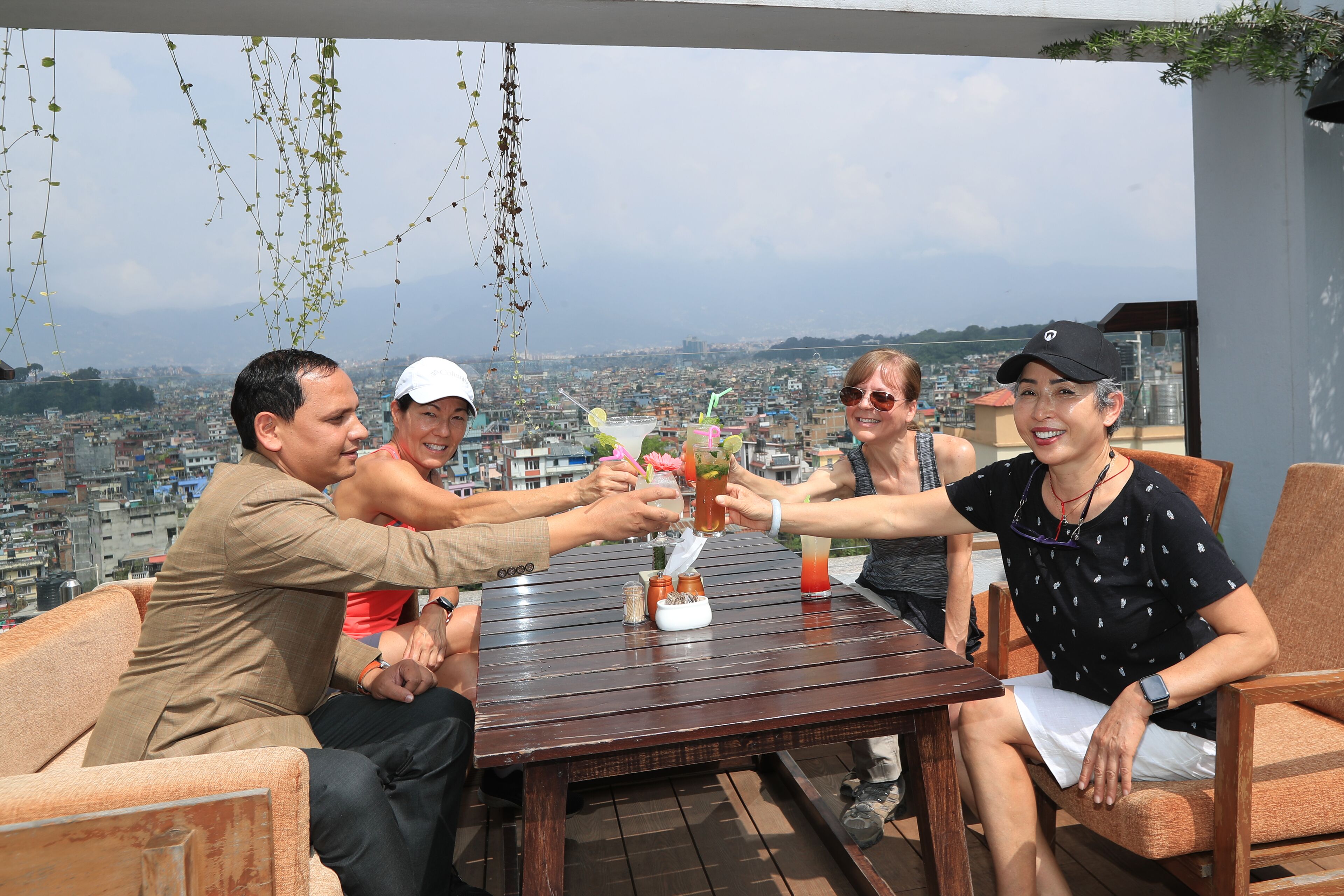 A group of people toasting with drinks at a wooden table with a city view in the background.