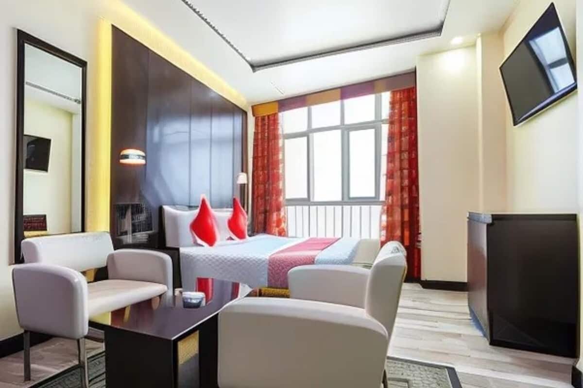 A hotel room with a bed, two white armchairs, a small table, a TV, and a window with curtains.