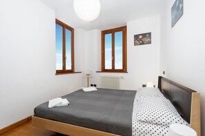 Ein Schlafzimmer mit einem Bett, zwei Fenstern, einem Heizkörper und einem Wandbild.