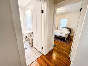 Een gang die leidt naar een slaapkamer met een bed, nachtkastje en raam. Een badkamer met een wastafel en spiegel is zichtbaar door een geopende deur.