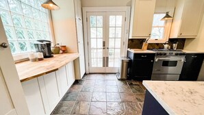 Une cuisine avec un sol carrelé en pierre, des meubles blancs, un plan de travail en bois et une vue sur l’extérieur grâce à une porte vitrée.
