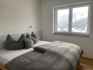 Quarto com cama, almofadas cinzentas, abajur e janela com vista para as montanhas.
