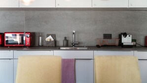 A modern kitchen with a grey backsplash, stainless steel sink, and a red microwave. There are two chairs with a purple towel draped over one.
