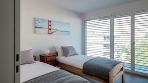 A bedroom with two beds, a painting of the Golden Gate Bridge, a nightstand, and a large window with blinds.