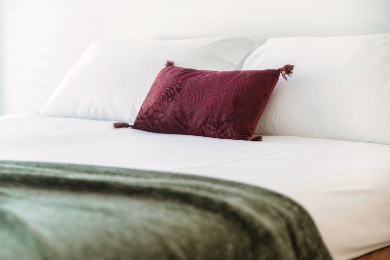 Una cama con s ábanas blancas y una almohada color burdeos.