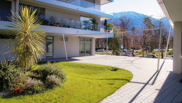 A modern multi-story building with a covered outdoor area, a well-maintained lawn, and a parking lot with cars.