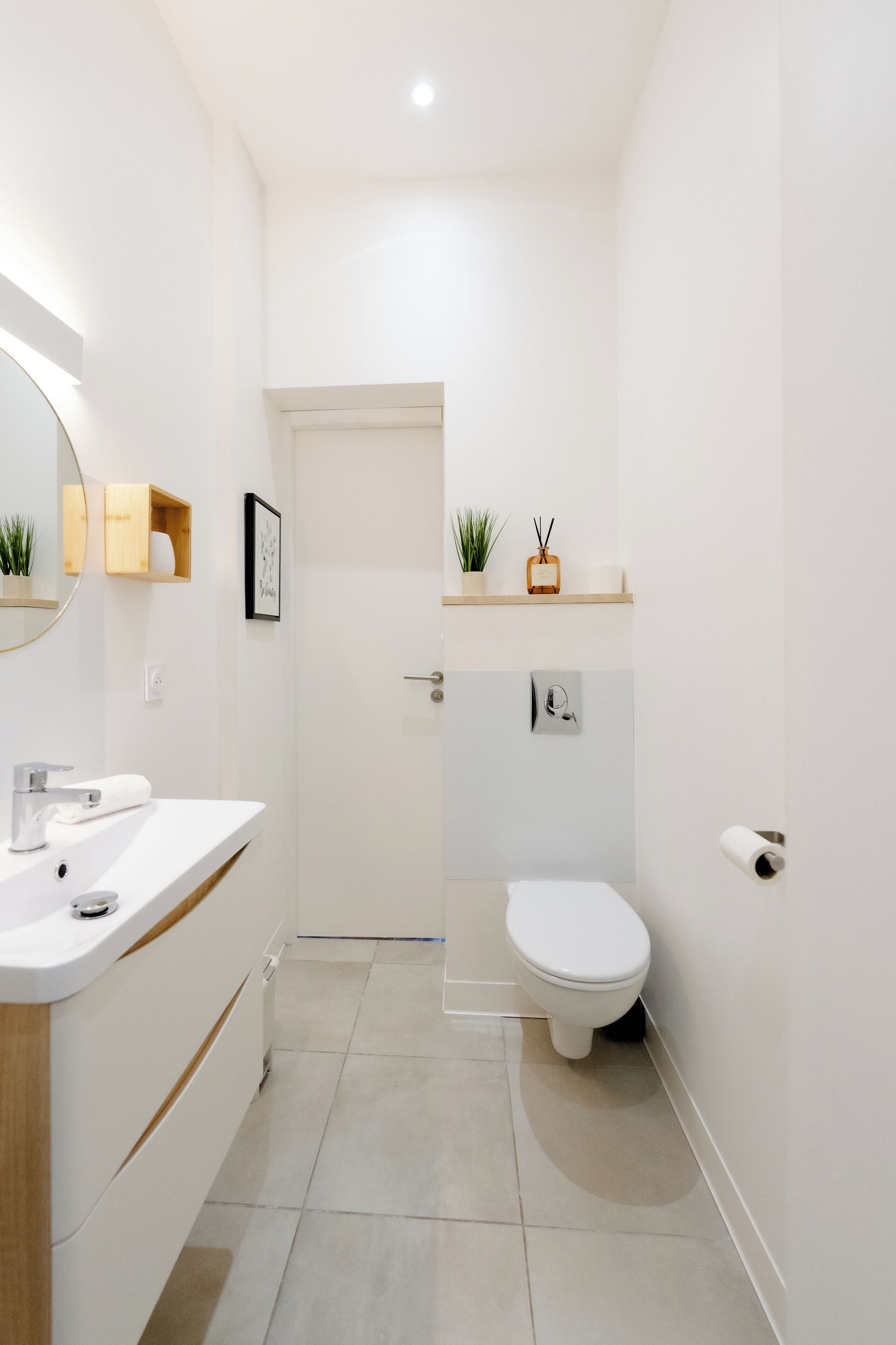 Ein modernes Badezimmer mit Waschbecken, Spiegel und Toilette in Weiß. Auf dem Regal stehen Pflanzen und an der Wand hängt ein gerahmtes Bild.