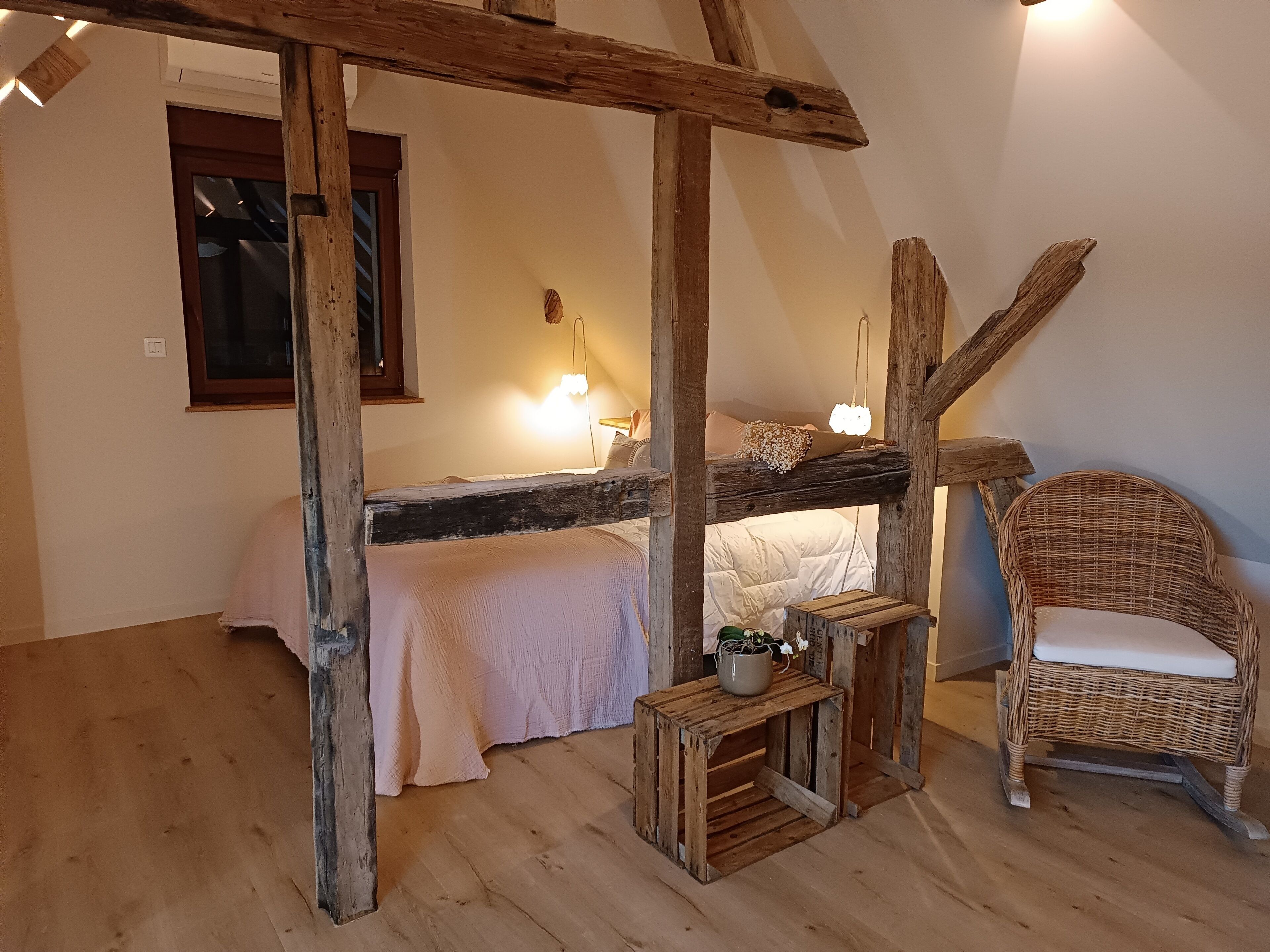 Een rustieke slaapkamer met zichtbare houten balken, een enkel bed, een rieten stoel en een klein houten tafeltje met een plantje.