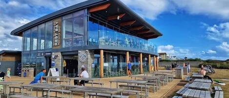A modern building with a glass facade, outdoor seating area, and a clear sky.