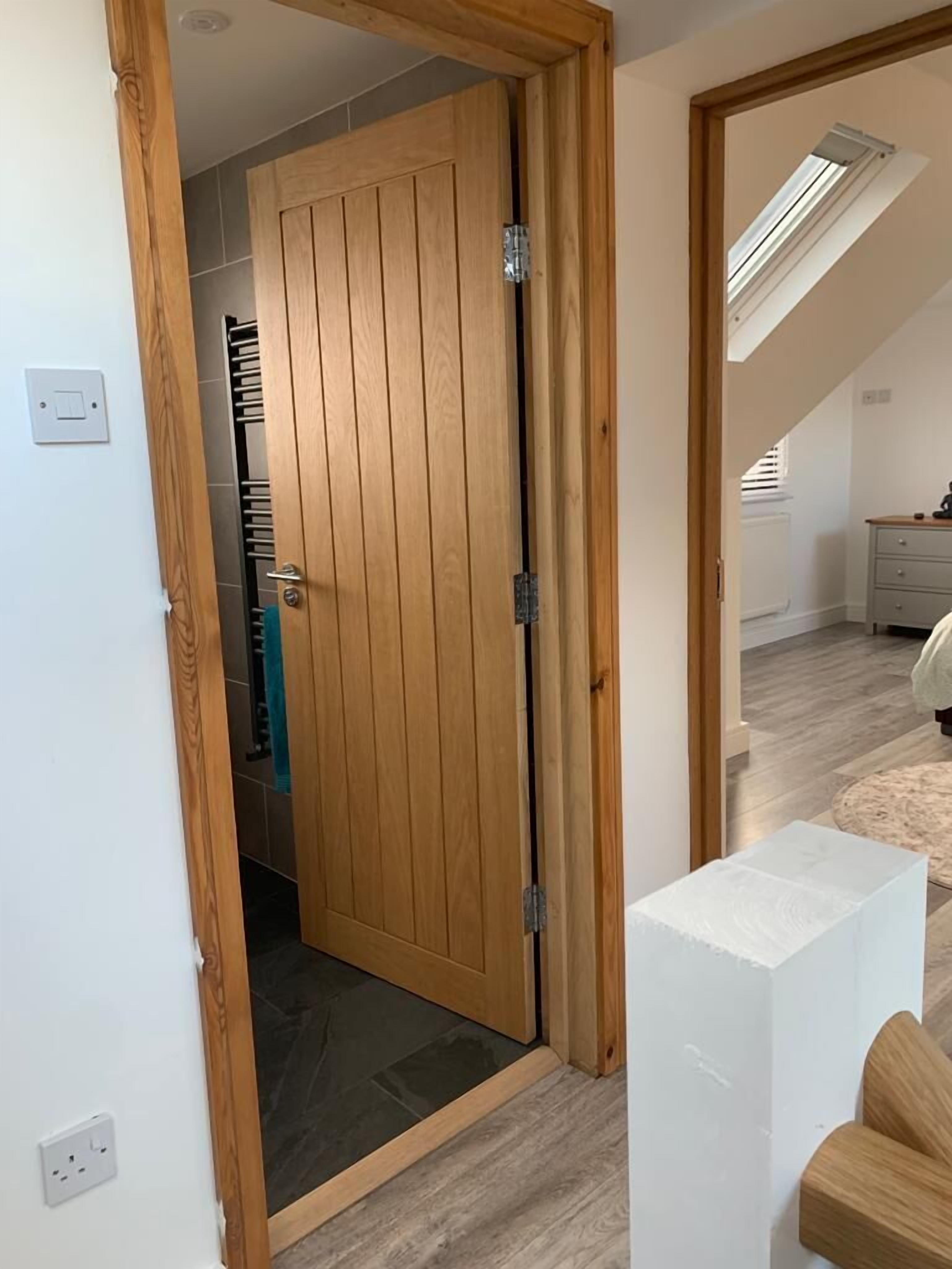 A room with a wooden door leading to a bathroom, a small white table, and a view of a bedroom with a bed and a window.