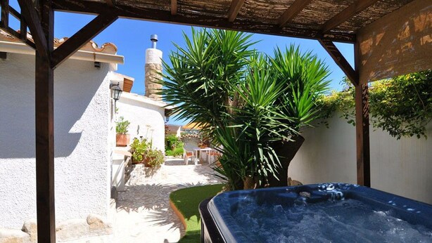 A hot tub in a covered outdoor area with a stone pathway, surrounded by greenery and a building with white walls. - Ina's vacation home (Teulada)