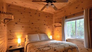 Une chambre à coucher dans une cabane en bois, avec un lit, des lampes de chevet, un ventilateur de plafond et une fenêtre avec des rideaux.