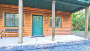 Une cabane en rondins avec une porte verte, deux fenêtres avec volets, un banc à l’extérieur et une allée gravillonnée.
