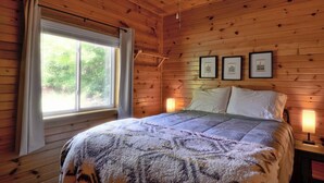 Une chambre de style cabane en bois, avec un lit, un ventilateur de plafond, une fenêtre avec des rideaux et des tableaux encadrés accrochés au mur.
