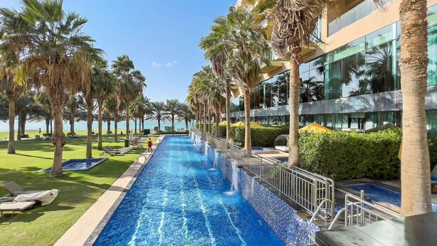 A swimming pool with a view of the sea, surrounded by palm trees and a modern building. - WelHome - Deluxe Apartment With Panoramic Sea Views (Dubai)