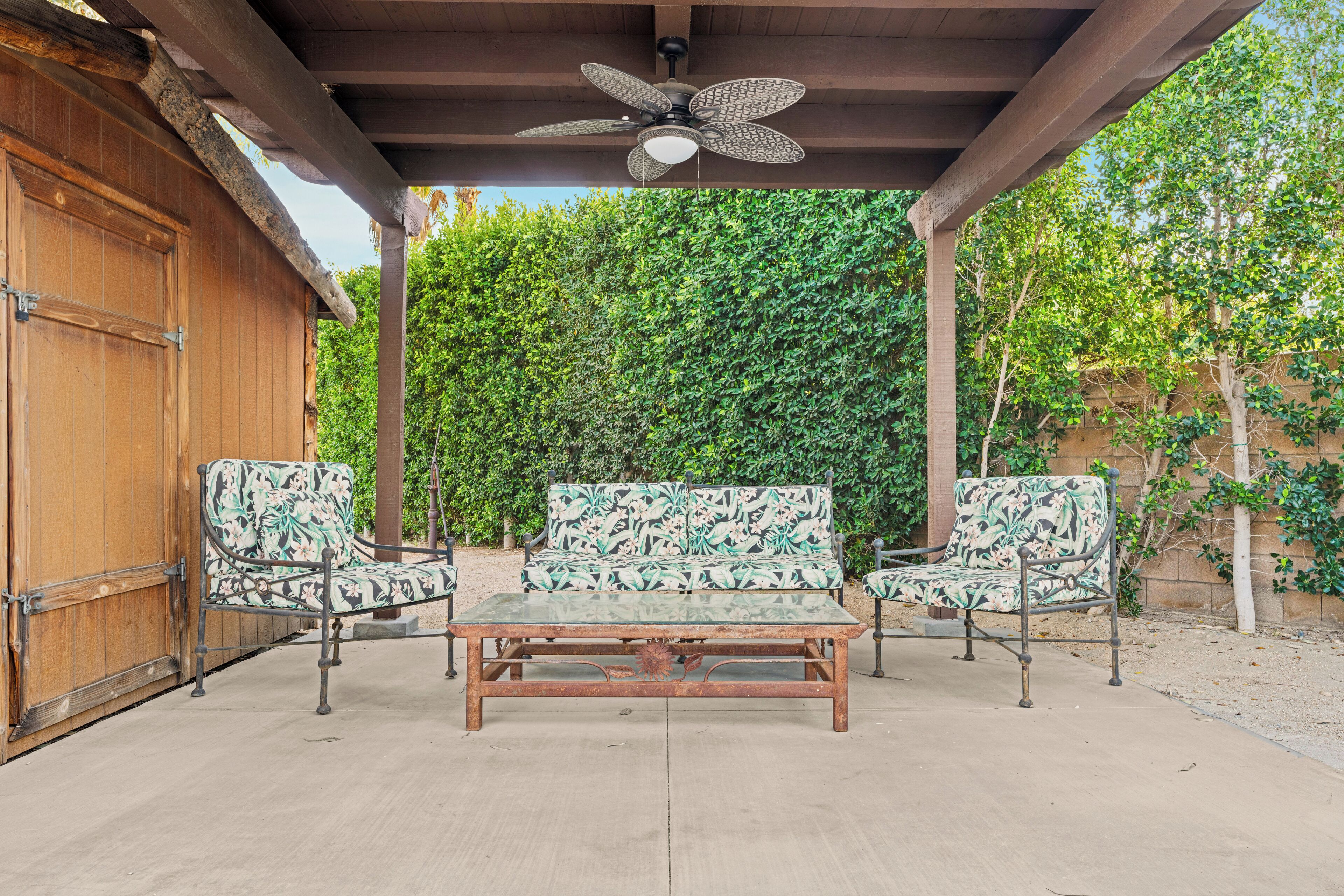 Eine Terrasse im Freien mit einem Holztisch und zwei gemusterten Stühlen, einem Deckenventilator und üppigem Grün im Hintergrund.