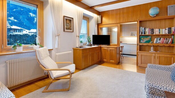 Een moderne woonkamer met houten vloer, een witte radiator, een witte stoel, een bank, een televisie, een boekenkast en een raam met uitzicht op besneeuwde huizen.