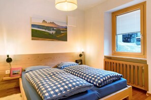 Un dormitorio con cabecera de madera, una ventana con persianas, una cama con ropa de cama a cuadros, una mesita de noche con un libro y una lámpara de pared.