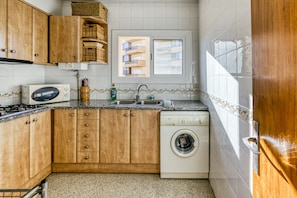 A compact kitchen with wooden cabinets, a washing machine, a microwave, and a window with a view of a building.