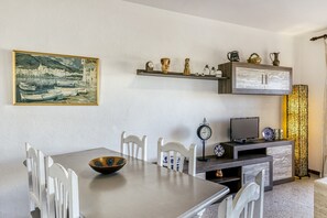 A dining area with a table, chairs, a bowl, a painting, a shelf with decorative items, a television, and a tall, textured lamp.