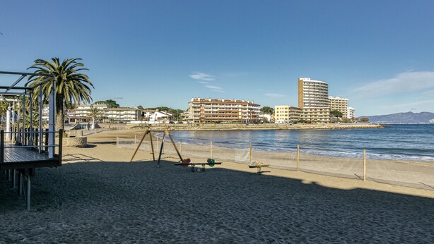 A beach with a playground, a palm tree, and buildings in the background. - Apartment 'Sol Y Mar 2 Dorm Playa Riells' with Balcony (L'Escala)