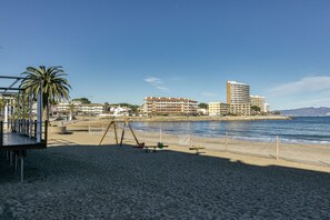 A beach with a playground, a palm tree, and buildings in the background. - Apartment 'Sol Y Mar 2 Dorm Playa Riells' with Balcony (L'Escala)