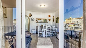 A living room with a sofa, a dining table with chairs, a coffee table, and a view of the city through a window. - Apartment 'Sol Y Mar 2 Dorm Playa Riells' with Balcony (L'Escala)