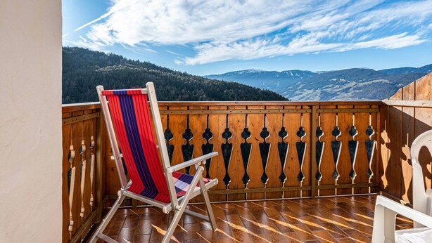 Un balcon en bois avec une chaise à rayures rouges et bleues, une chaise blanche et une vue sur des montagnes lointaines.