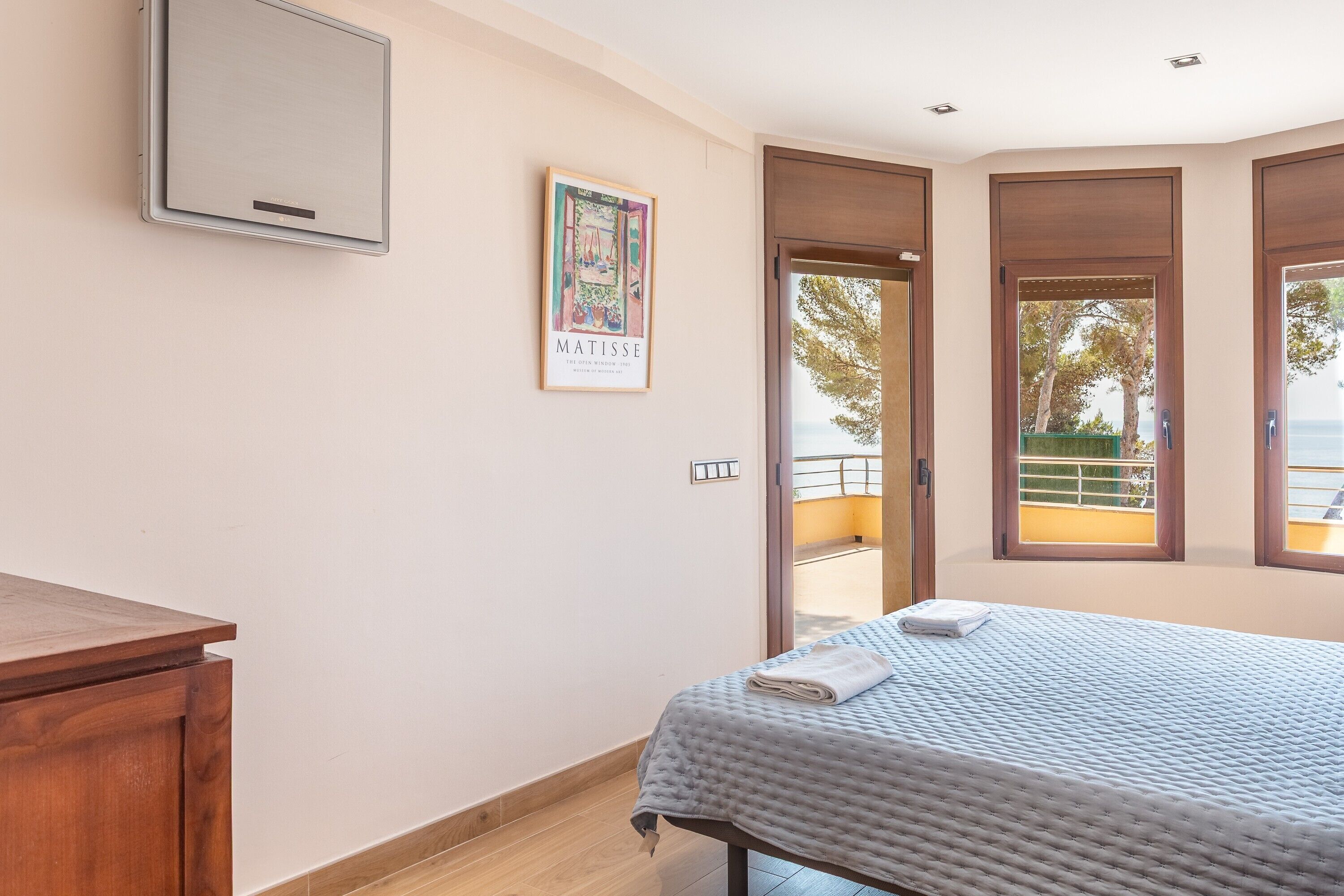 Une chambre à coucher avec un lit, un bureau en bois, une œuvre d’art encadrée et une fenêtre donnant sur un paysage.