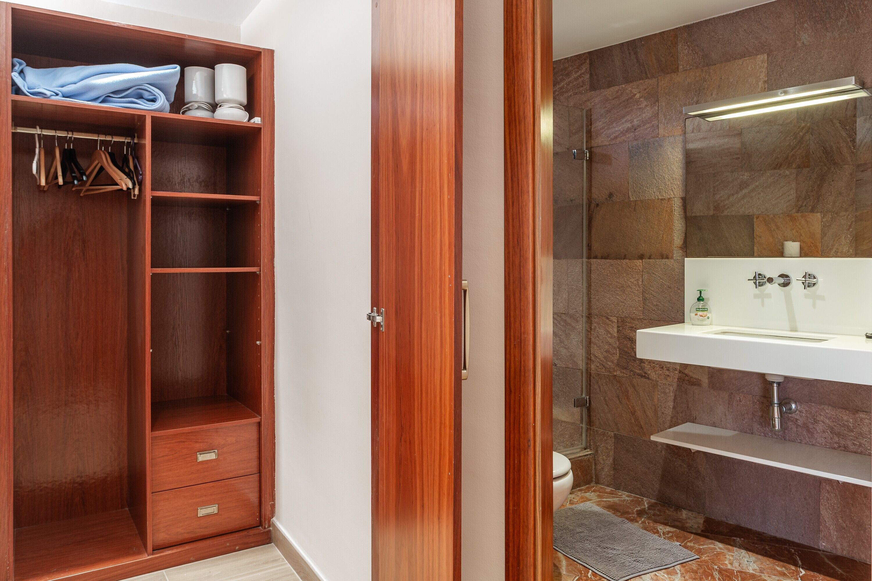A wooden wardrobe with hanging space, a mirror, and a bathroom with a marble floor and a sink.