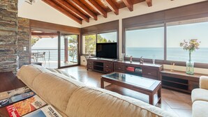 Ein geräumiges Wohnzimmer mit Steinwand, Holzbalkendecke, einem großen Fernseher und Blick auf das Meer.
