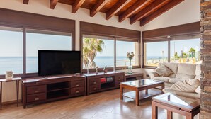 Ein Wohnzimmer mit Fernseher, Holzmöbeln, großen Fenstern mit Meerblick und einer Steinwand.