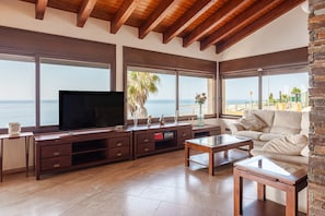 A living room with a TV, wooden furniture, large windows with a view of the sea, and a stone wall. - Villa 'Villa Con Infinity Pool' with Sea View, Wi-Fi and Air Conditioning (Lloret de mar)