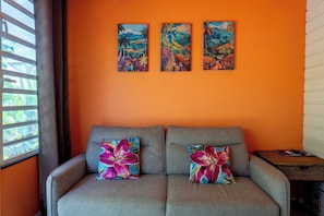 Ein Zimmer mit einer orangefarbenen Wand, einem grauen Sofa mit floralen Kissen und drei Bildern darüber.