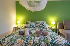 Ein ordentlich gemachtes Bett mit Bettdecke mit tropischem Blattmuster, zwei Nachttische mit Lampen, ein Baldachin und ein Stuhl.