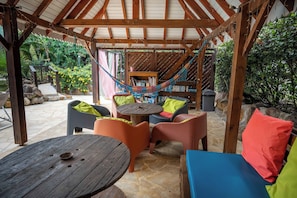 A covered outdoor seating area with a hammock, wooden tables, and colorful chairs. - 900m from the beach, the Bagatelle gites ** 2/4 pers, swimming pool and SPA (Sainte-Rose)