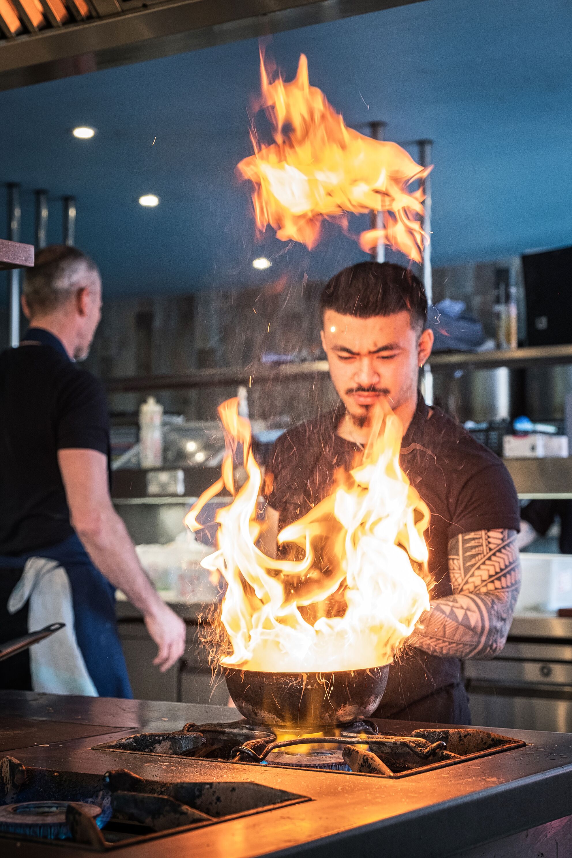 A chef performing a flambé with a flaming pan on a stove in a kitchen.