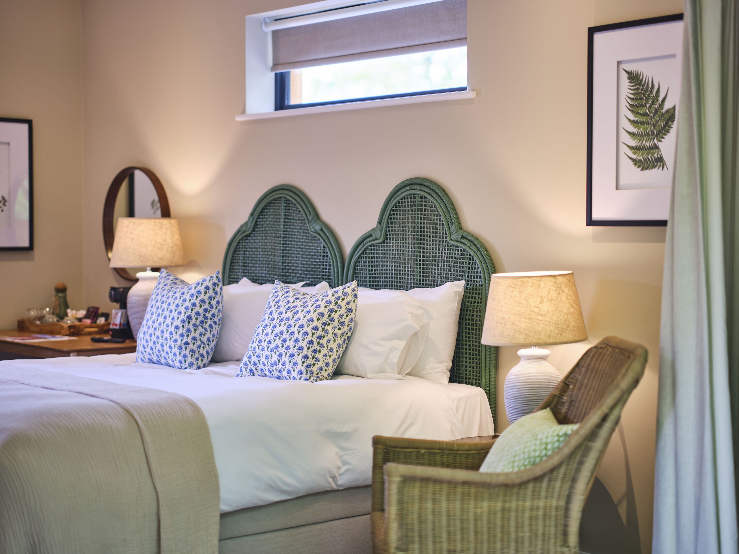 A neatly made bed with patterned pillows, a green headboard, a bedside table with a lamp, and a framed plant on the wall.